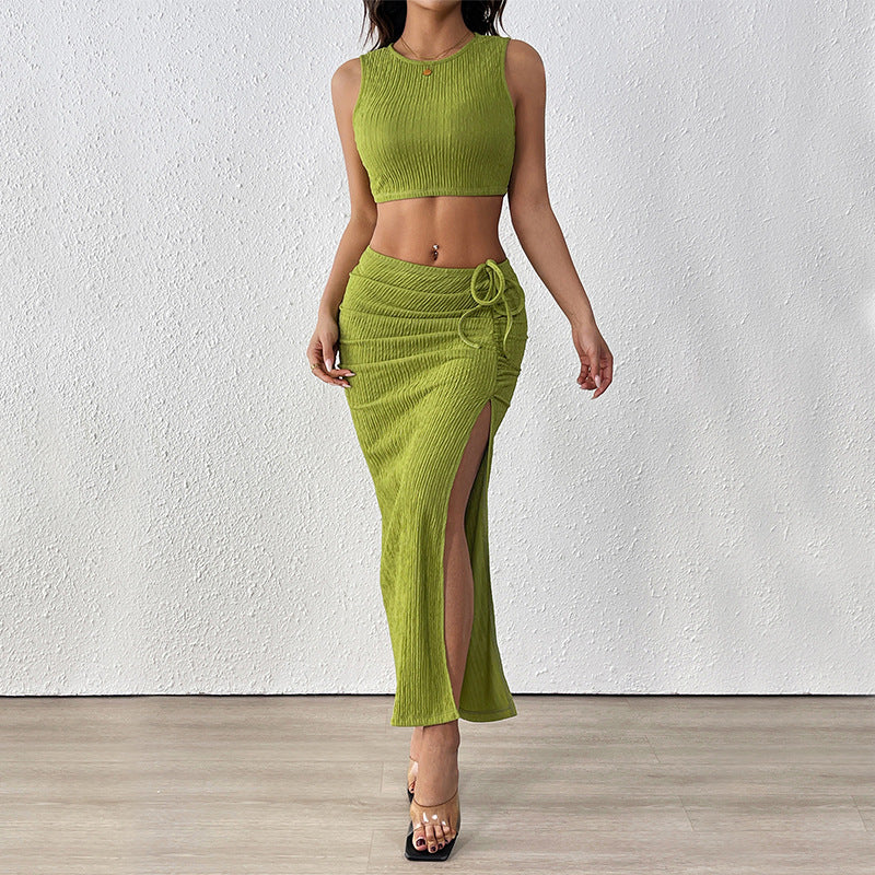 A woman wearing a green two-piece set featuring a ribbed crop top and a matching high-waisted skirt with a ruched side tie and a thigh-high slit, paired with clear high heels.