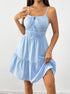 A woman is wearing a powder blue tiered sundress with tie-front neckline and smocked bodice, posing with one hand touching the fabric