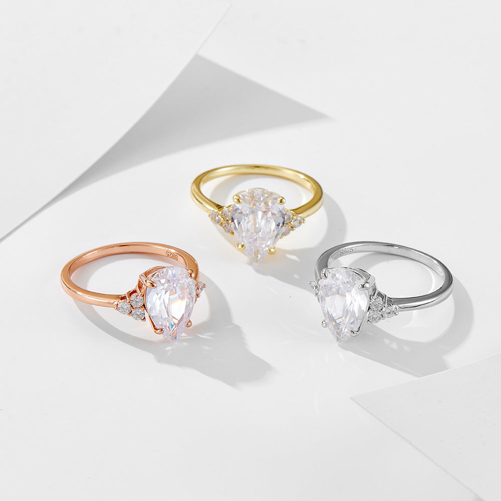 Three teardrop engagement rings in rose gold, yellow gold, and silver displayed on white geometric surface, each featuring pear-cut crystals