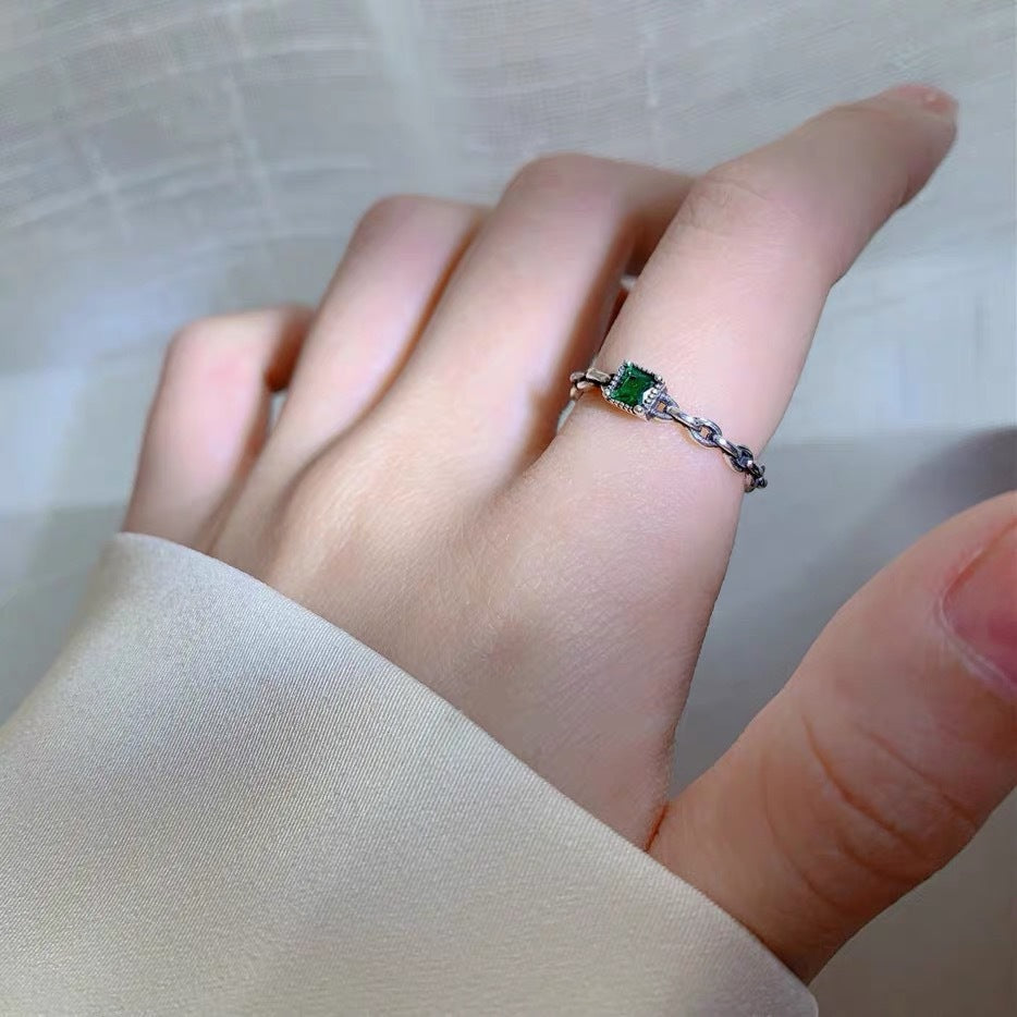 A woman wearing silver chain ring with emerald green stone on finger, hand against white fabric with soft natural lighting