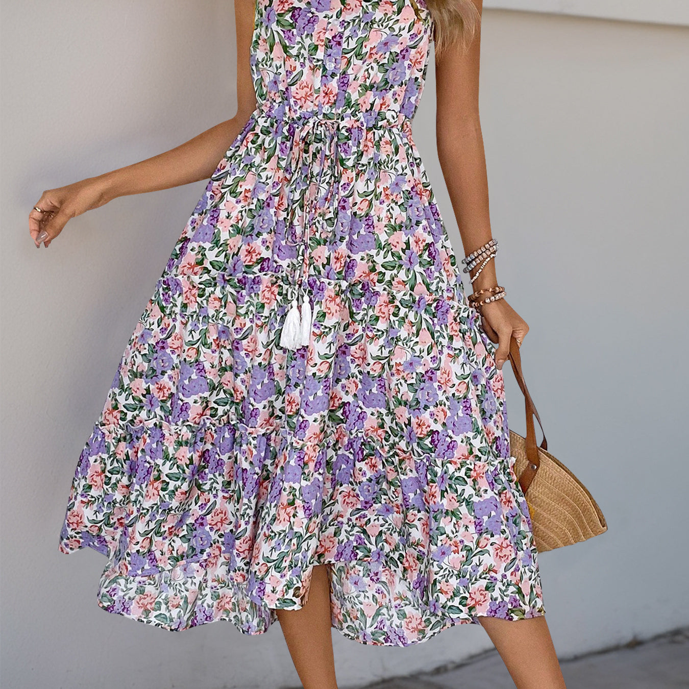 A woman wearing a floral midi dress with spaghetti straps, a drawstring waist, and tiered ruffles. She carries a woven handbag and wears strappy sandals.