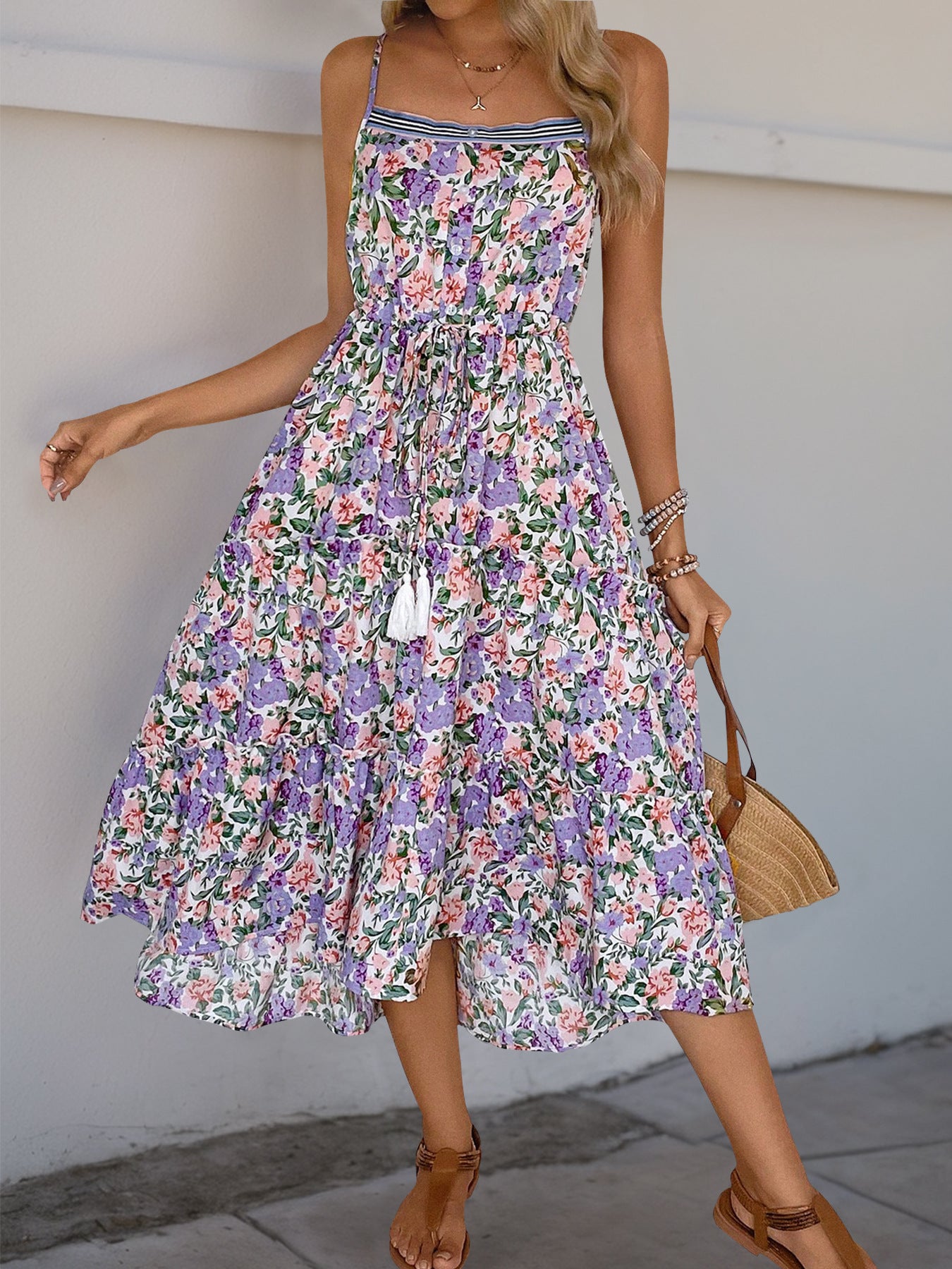 A woman wearing a floral midi dress with spaghetti straps, a drawstring waist, and tiered ruffles. She carries a woven handbag and wears strappy sandals.
