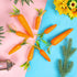  A set of artificial carrot decorations with green leaves, placed on a split pink and blue background with greenery around.