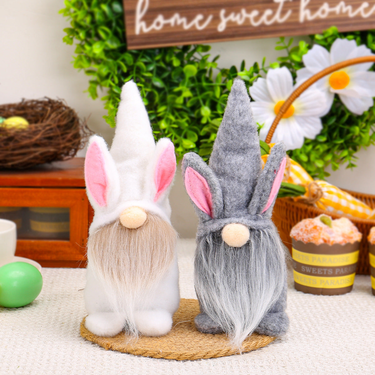 A pair of Easter gnome plush toys with bunny ears, one in white and the other in gray, displayed on a table with festive decorations.