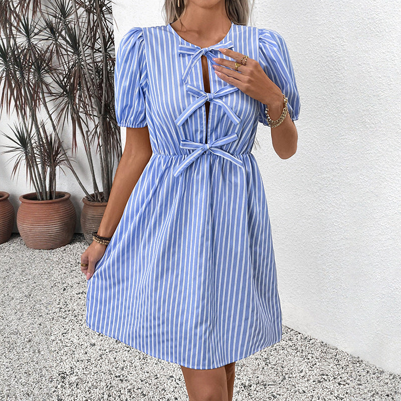 A woman wears a blue and white striped dress with short puff sleeves and three bow ties on the front, standing against a white wall.