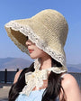 Woman wearing a beige crocheted sun hat with lace trim and a tie, standing outdoors against a blue sky.