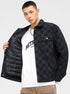 A model is showing the quilted lining of a black checkered jacket over white graphic tee and tan pants against white backdrop