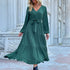A woman is posing in an emerald green tiered maxi dress with v-neck, button front, long balloon sleeves and waist tie, styled with layered necklaces
