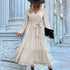 A woman is posing in a cream tiered maxi dress with button details, self-tie belt, bishop sleeves and ruffle hem, accessorized with black hat