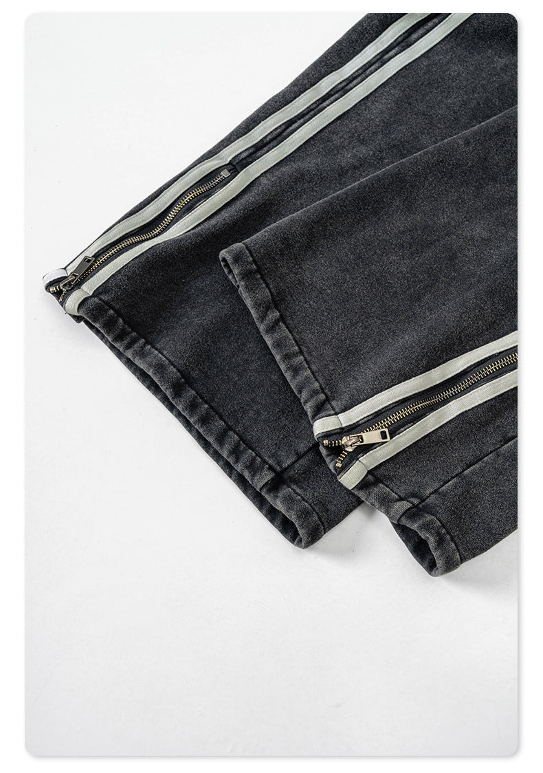 Detail of ankle zipper with gray tape trim on black denim track pants, showing quality construction and hardware