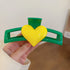  A glossy green hair clip with a bold yellow heart centerpiece, held in a person’s hand against a blurred background.