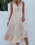 A woman is wearing a cream tiered maxi dress with button front detail, standing against a white stucco wall with greenery