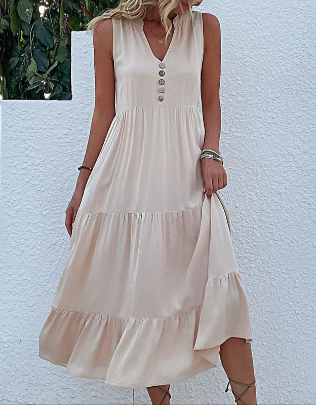 A woman is wearing a cream tiered maxi dress with button front detail, standing against a white stucco wall with greenery