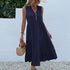 A woman is wearing a navy sleeveless tiered dress with button details, carrying a straw handbag against a textured white backdrop