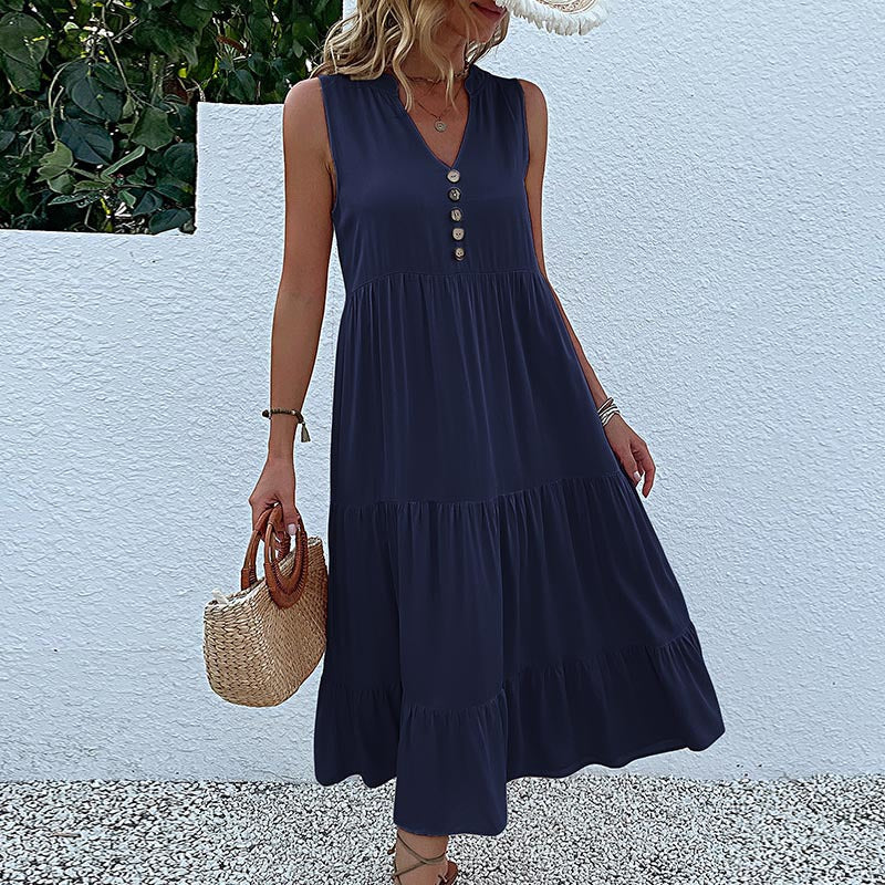 A woman is wearing a navy sleeveless tiered dress with button details, carrying a straw handbag against a textured white backdrop