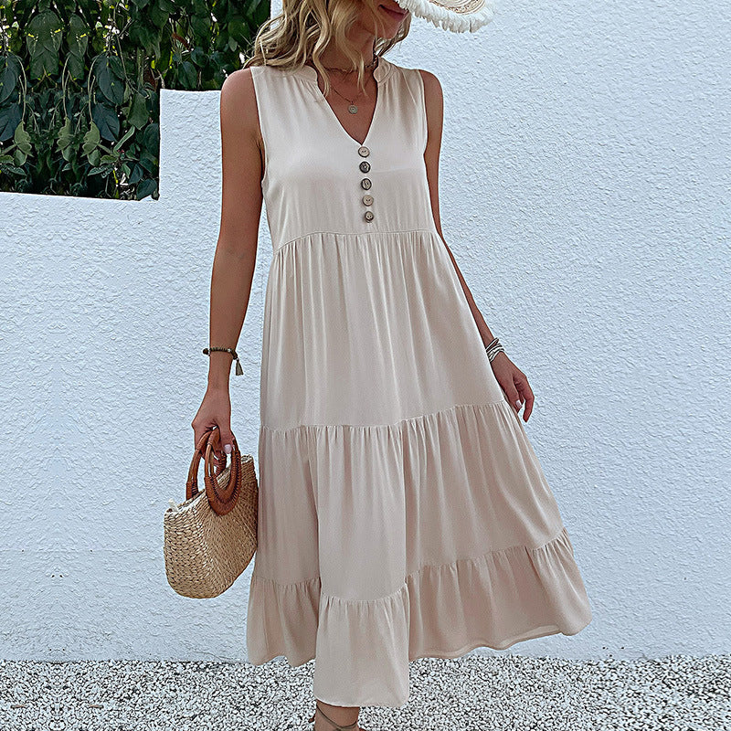 A woman is wearing a cream v-neck tiered midi dress, holding a straw bag against a white wall with lush greenery visible