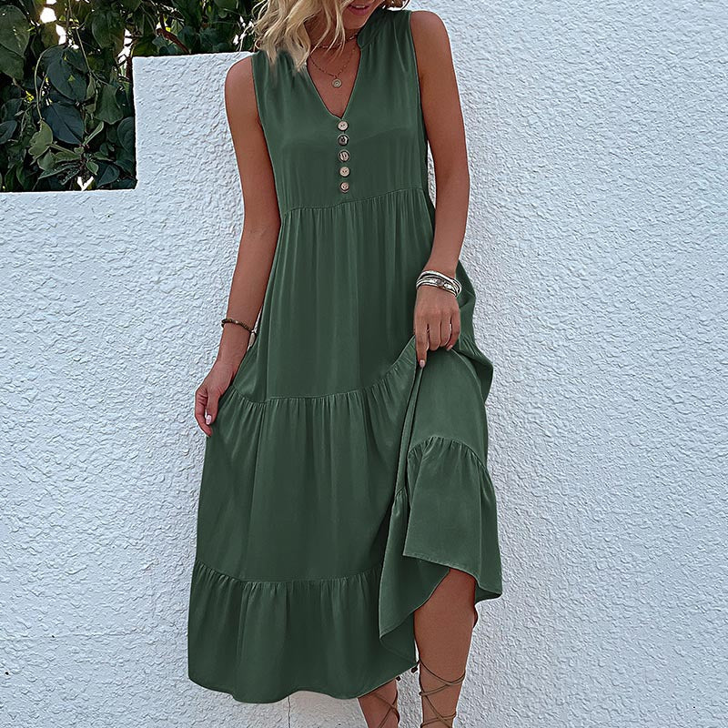 A woman is wearing an olive green sleeveless tiered dress with button front detail, lifting the fabric against a white wall