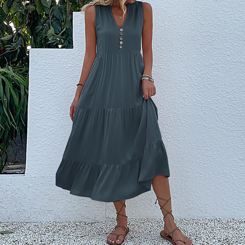 A woman is wearing a slate blue v-neck tiered dress with decorative buttons, holding a straw bag against a white backdrop.