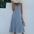 A woman is wearing a dusty blue tiered dress with front buttons, holding a straw hat that covers her face against a white wall