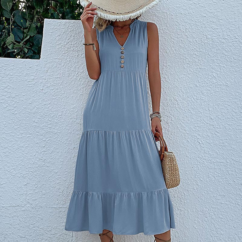 A woman is wearing a dusty blue tiered dress with front buttons, holding a straw hat that covers her face against a white wall