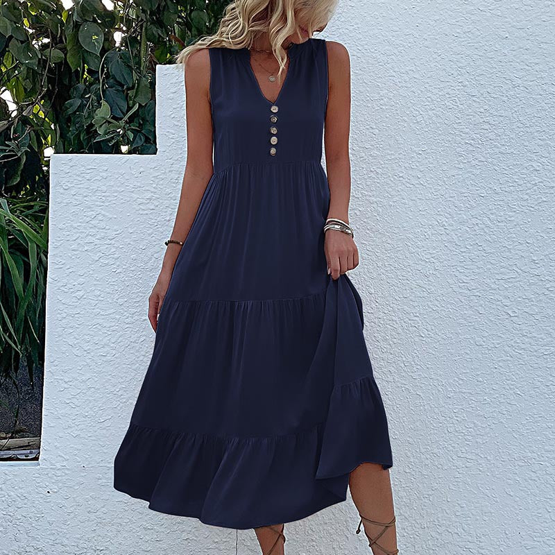 A woman is wearing a navy sleeveless v-neck dress with tiered skirt, standing against a white wall with patterned ground