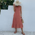 A woman is wearing a pink sleeveless midi dress with tiered design, holding a straw hat that partially covers her face