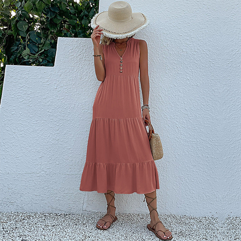 A woman is wearing a pink sleeveless midi dress with tiered design, holding a straw hat that partially covers her face