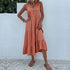 A woman is wearing a coral tiered midi dress with decorative buttons, standing with hand on hip against a white wall