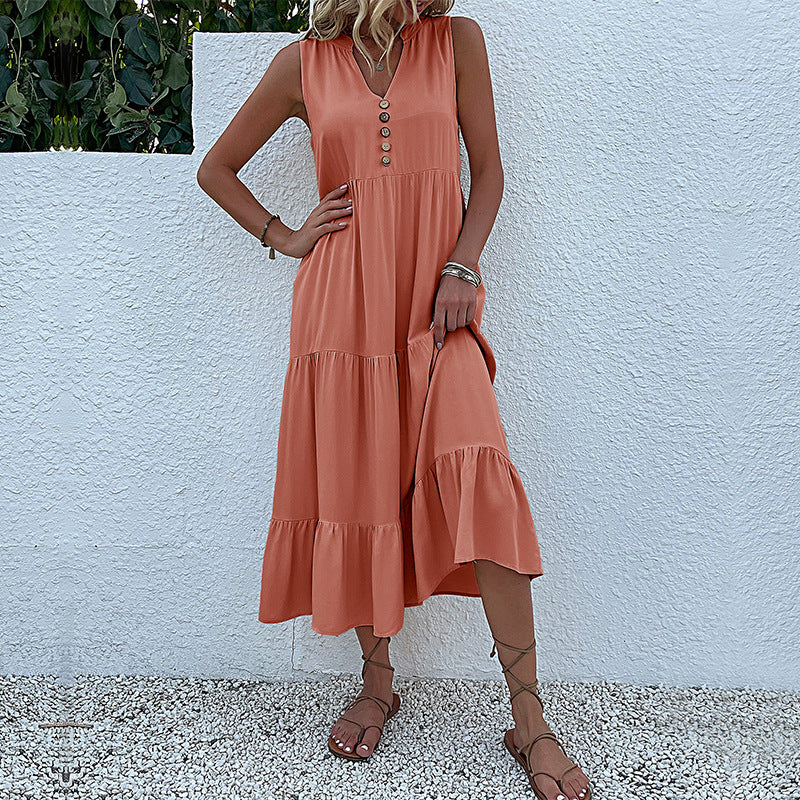A woman is wearing a coral tiered midi dress with decorative buttons, standing with hand on hip against a white wall
