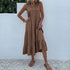 A woman is wearing a mocha tiered maxi dress with button details, standing against a white wall with visible greenery