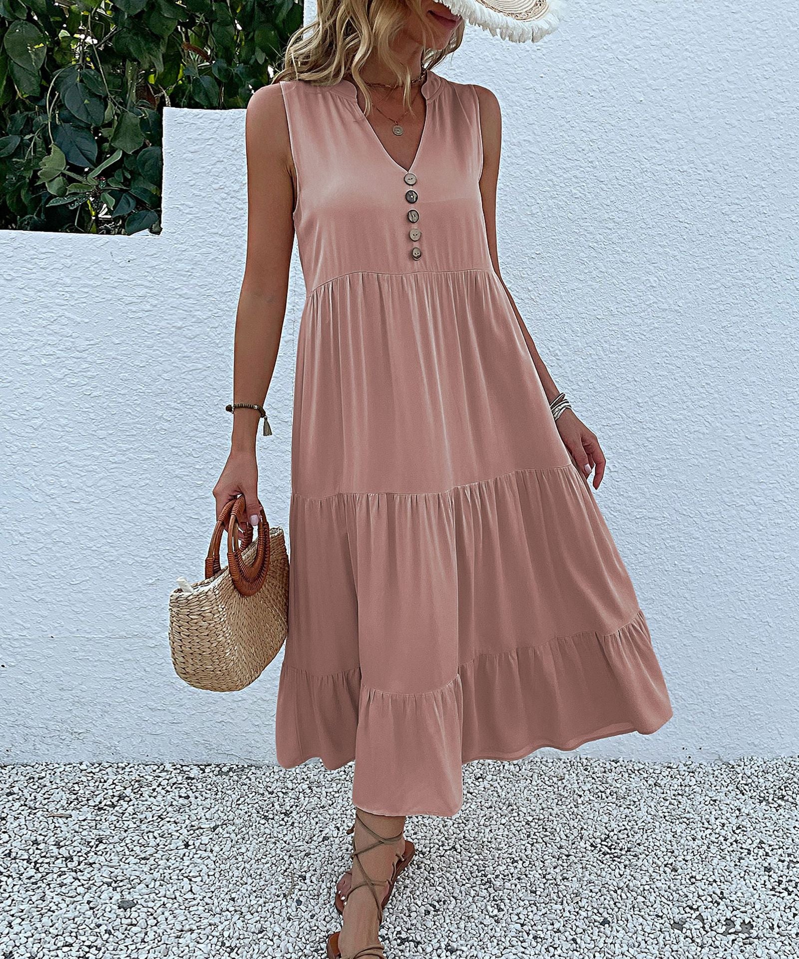 A woman is wearing a blush tiered midi dress with decorative front buttons, holding a straw basket against a white wall