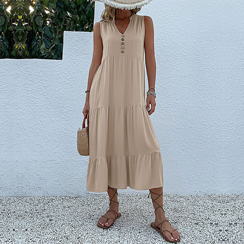 A woman is wearing a beige tiered maxi dress with button detail and v-neckline, carrying a small straw bag against white wall