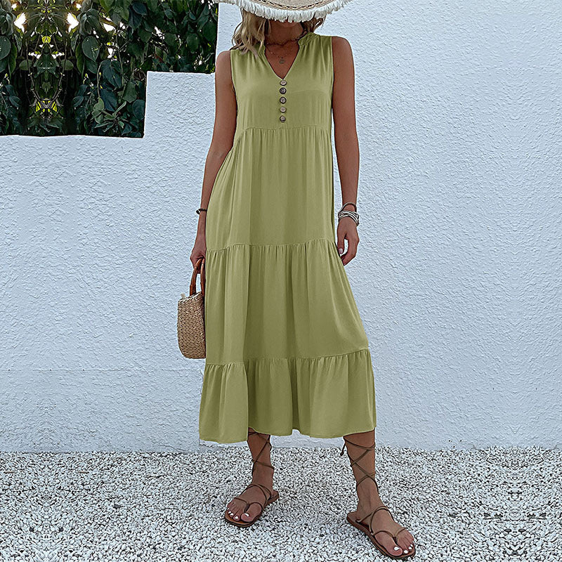 A woman is wearing a sage green tiered maxi dress with v-neck and button detail, carrying a straw bag against a white wall