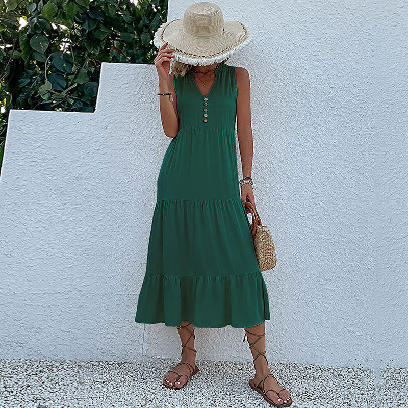 A woman is wearing a forest green tiered maxi dress with decorative buttons, holding a straw hat that covers her face