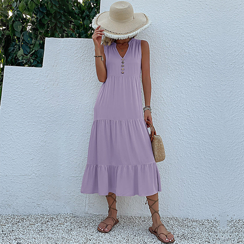 A woman is wearing a lavender sleeveless tiered dress with button front detail, carrying a straw bag against a white background