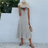 A woman is wearing a light gray tiered midi dress with v-neck and button detail, accessorized with a straw hat against white wall