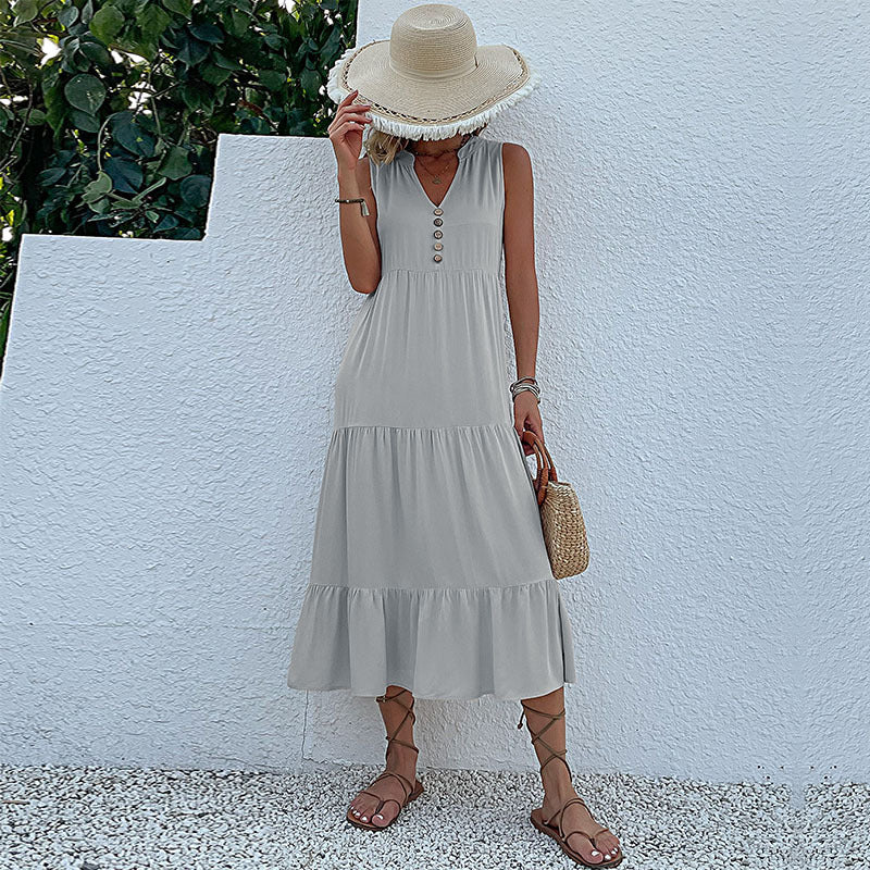 A woman is wearing a light gray tiered midi dress with v-neck and button detail, accessorized with a straw hat against white wall