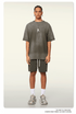 Model with braided hair wearing charcoal green t-shirt and matching shorts, silver pendant necklace, white socks against cream background