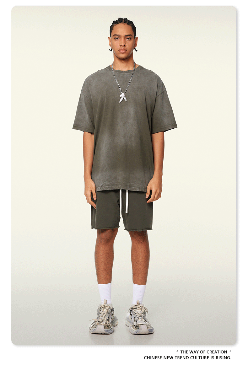 Model with braided hair wearing charcoal green t-shirt and matching shorts, silver pendant necklace, white socks against cream background