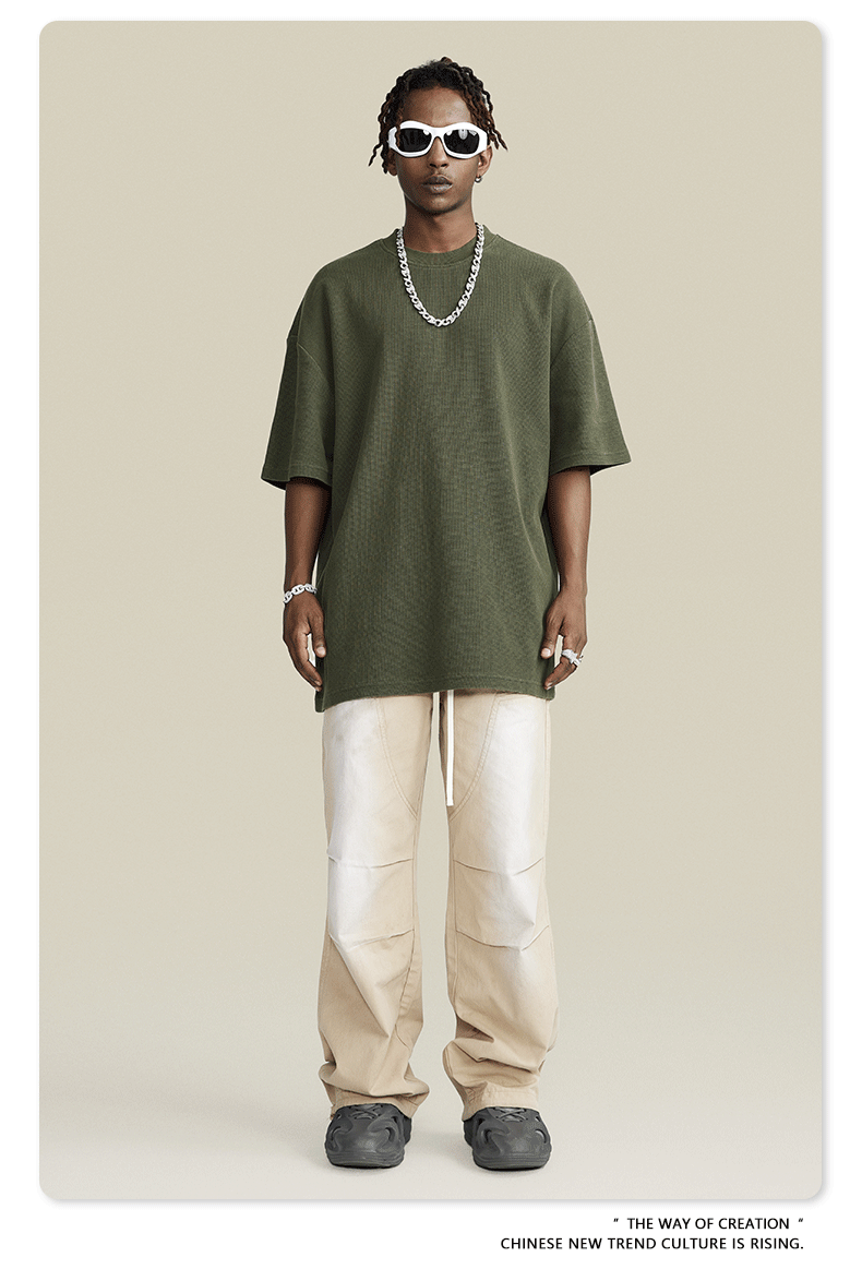 Model in olive t-shirt, white wide-leg pants, white sunglasses, silver chain against beige background