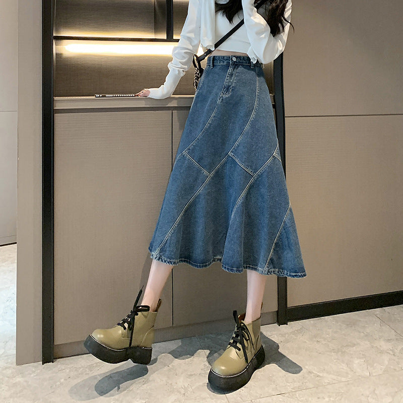 A woman is wearing a flared blue denim midi skirt with contrast stitching, a white cropped sweater, and chunky olive-green lace-up boots
