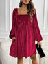 A woman wearing a burgundy ribbed velvet babydoll dress with balloon sleeves stands against white paneling, holding a black clutch