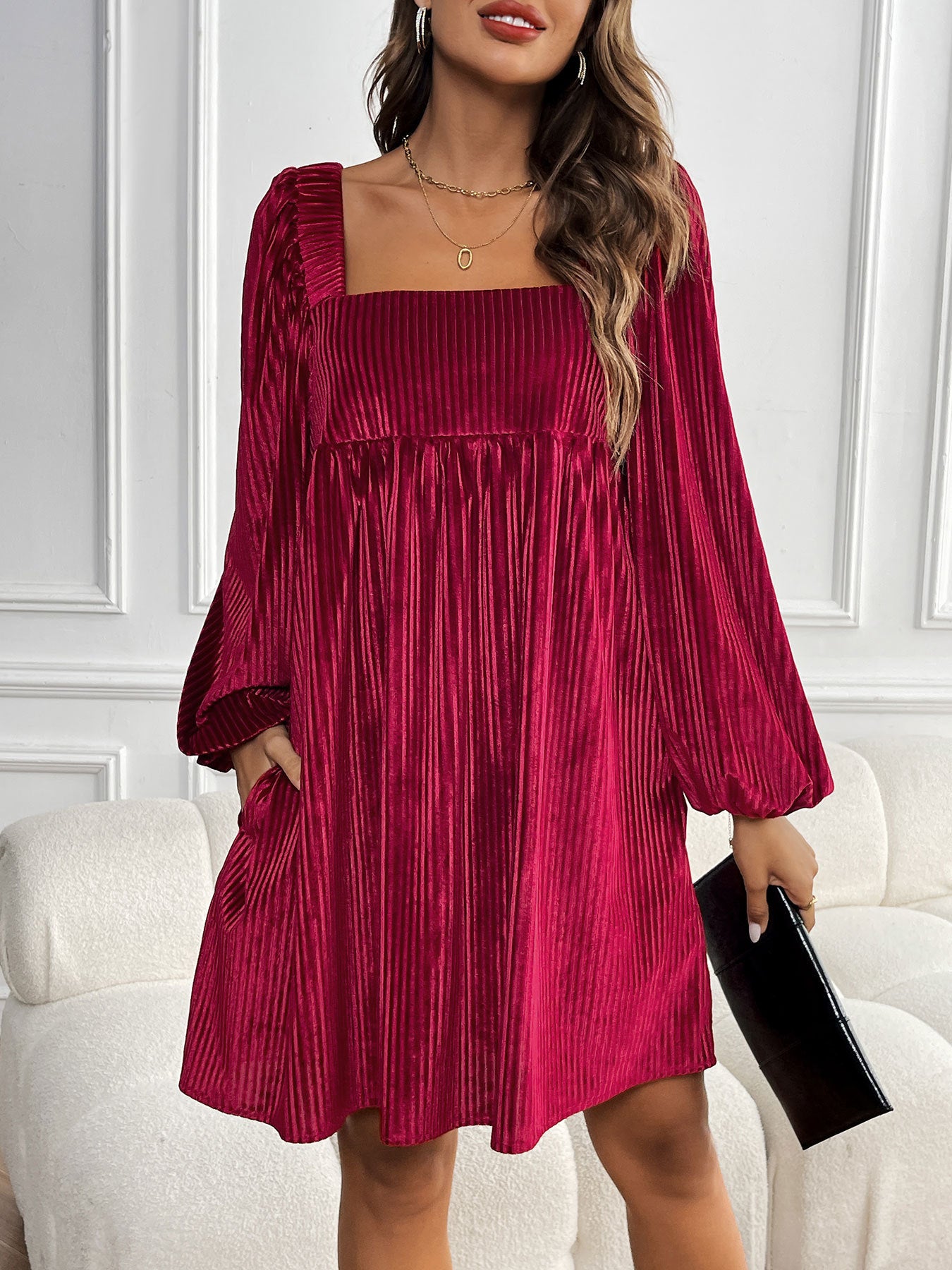 A woman wearing a burgundy ribbed velvet babydoll dress with balloon sleeves stands against white paneling, holding a black clutch
