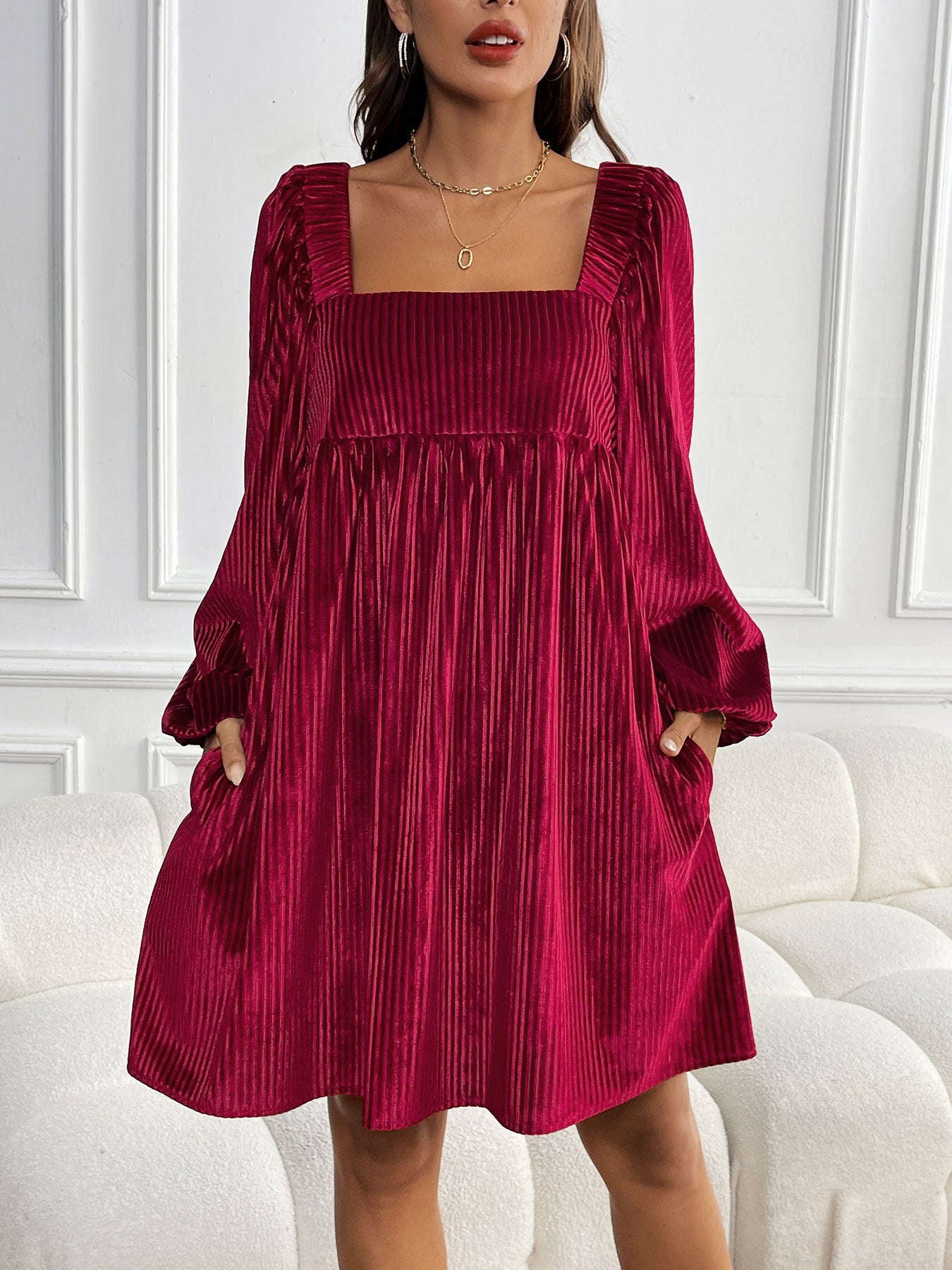 A woman wearing a burgundy pleated velvet mini dress with square neckline and balloon sleeves poses against white wall panels