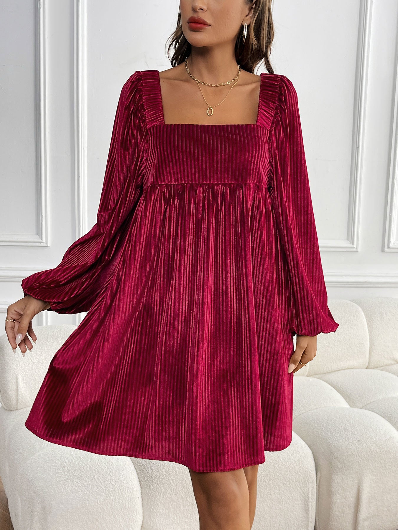 A woman in a burgundy velvet empire waist dress with puffy sleeves stands by a white sofa, showing the textured ribbed fabric