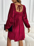 A woman wearing a burgundy velvet dress shows the back view with tie closure, standing next to a white couch with black clutch