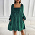 A woman wearing an emerald green ribbed velvet mini dress with square neckline and balloon sleeves poses by a white sofa