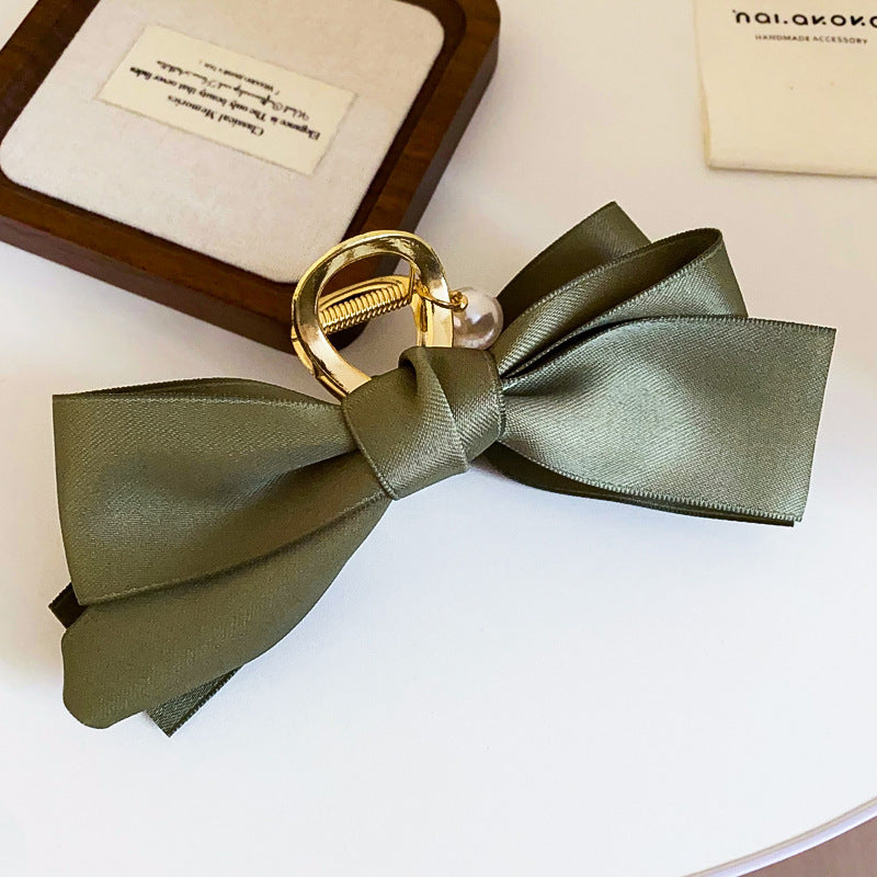 A dark green satin bow hair clip with a gold claw clasp and pearl detail, displayed on a white surface next to a wooden jewelry box.