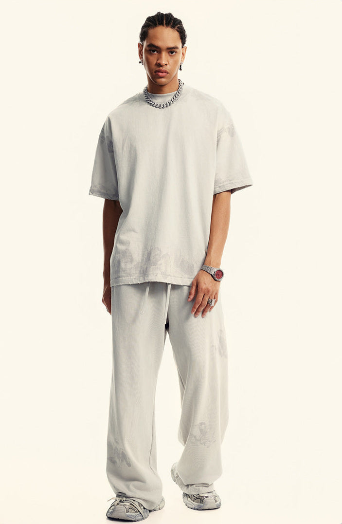 Front view of model in light beige oversized t-shirt and matching pants. Silver accessories include chain necklace and red-faced watch