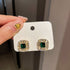 Square-cut emerald earrings with diamond halo settings displayed on white retail card showing luxury jewelry presentation